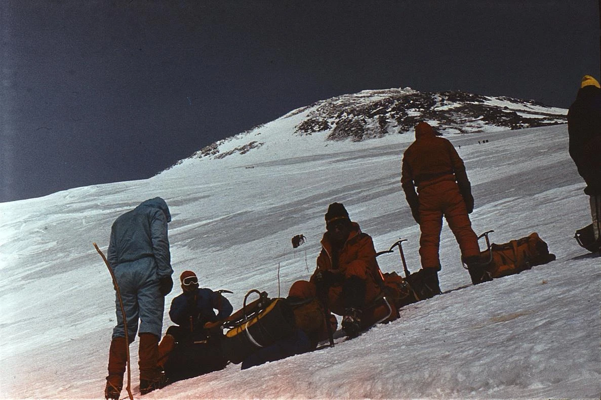Почивка по пътя – Йордан Зашев е седнал в средата с поглед към камерата; в далечината три групи напредват по ледника към върха (Кавказ ’83 – лагер-сбор за „Еверест ’84“)